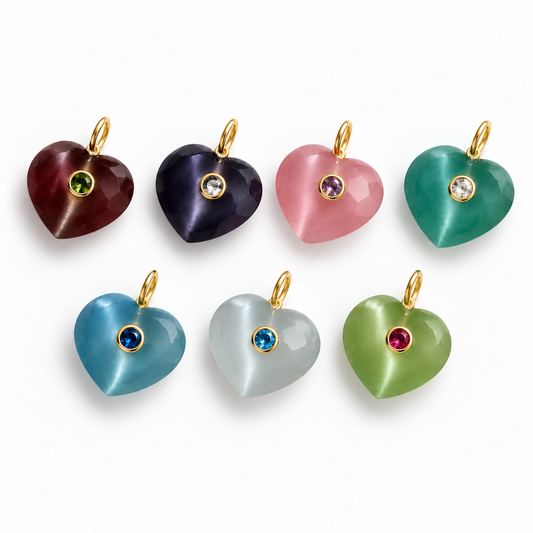 Seven faceted heart-shaped cat-eye jewelry pendants in various colors (maroon, purple, pink, emerald green, sky blue, milky white, and light green) with gold loops and round gemstone centers, arranged on a white background.