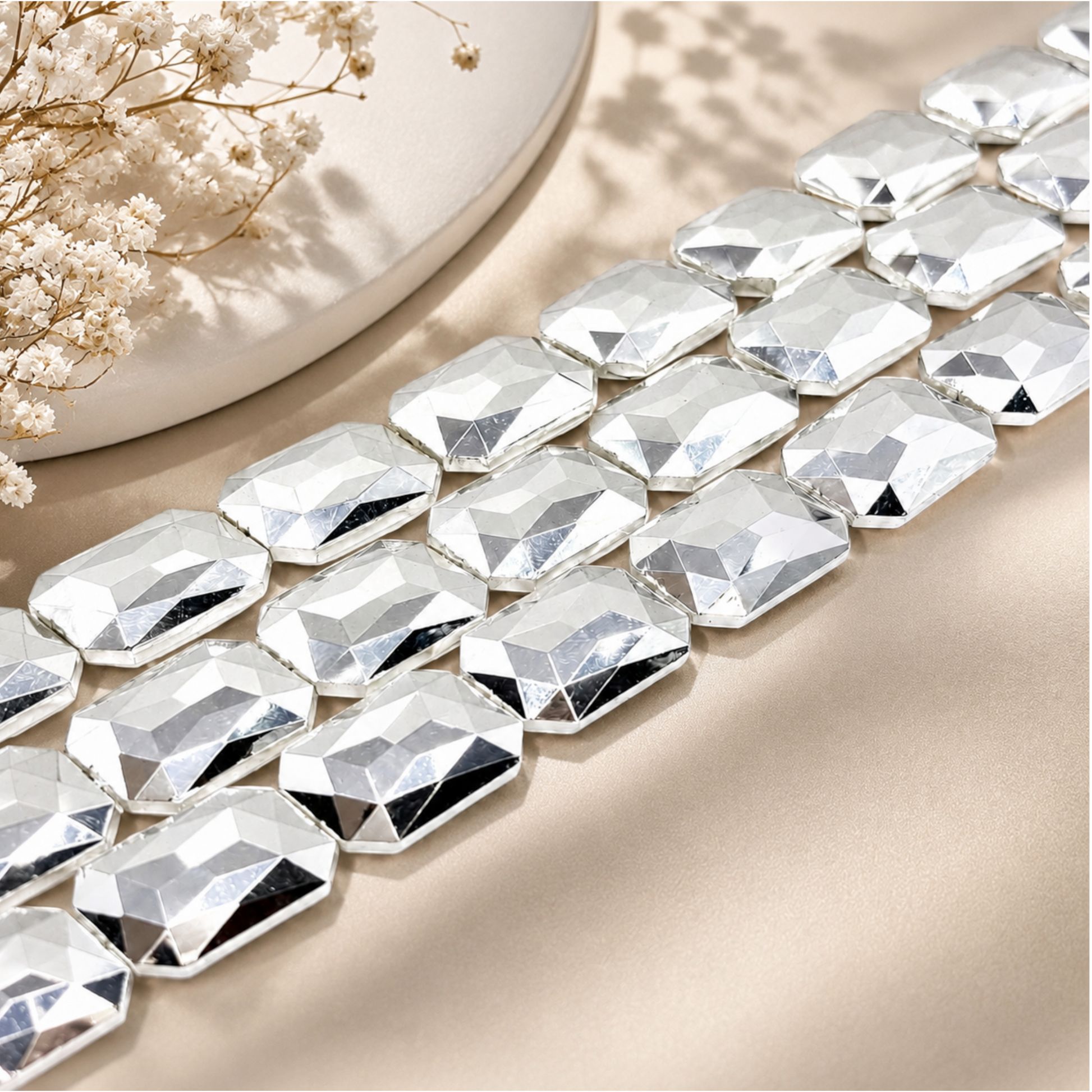 "Strands of large silver faceted crystal beads styled on a beige surface with dried white flowers and ceramic vase"